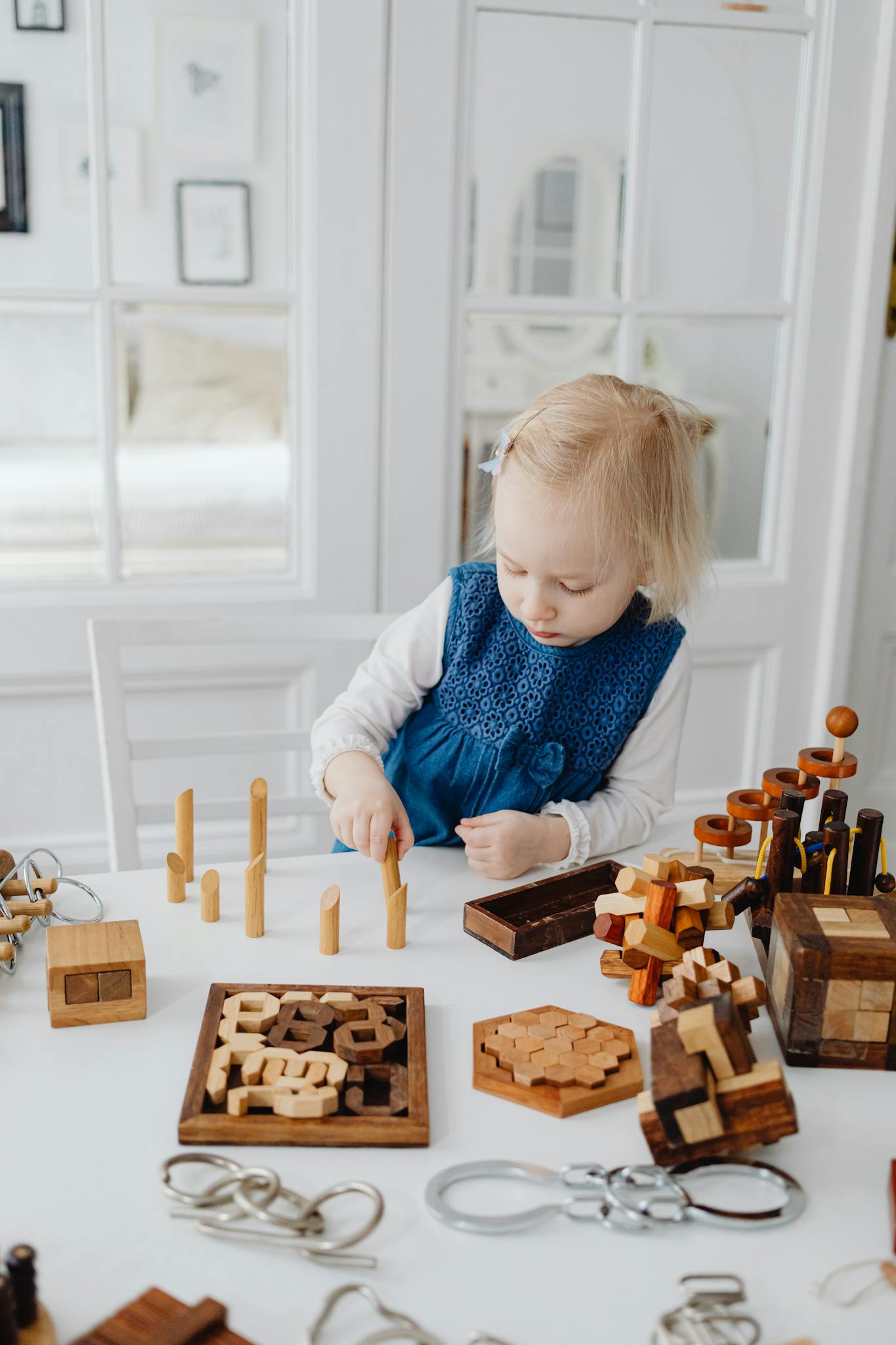 Child in blue dress playing with wooden puzzles at a table, enhancing cognitive skills indoors.